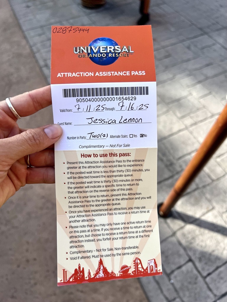 A hand holding a Universal Orlando Resort Attraction Assistance Pass. The pass has an orange header with the Universal globe logo and the words "Attraction Assistance Pass." Below is a barcode and details filled in by hand.