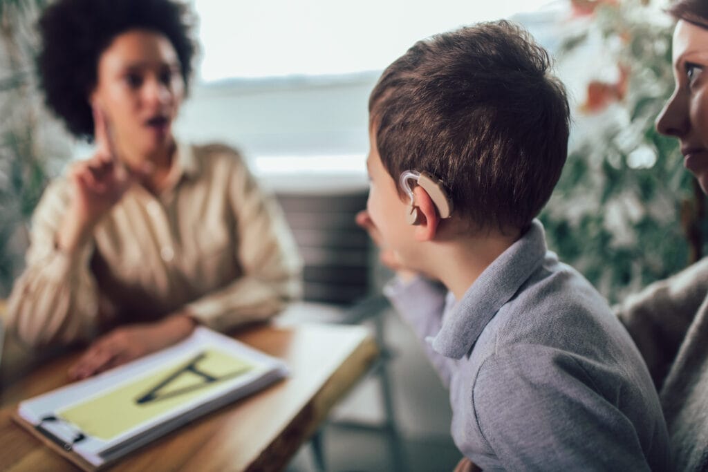 A young boy wearing a hearing aid sits at a table and gestures while facing a teacher who is signing. An adult sits beside him in a bright educational setting, suggesting a language or communication learning session.